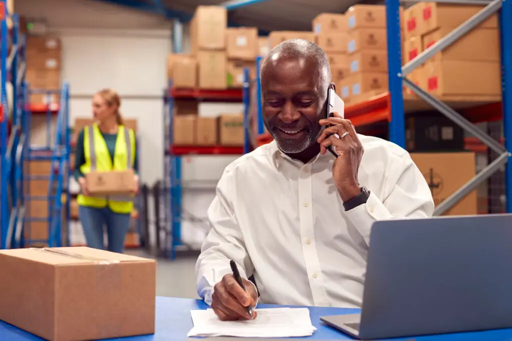 Male manager working on a laptop and talking on a mobile phone in a warehouse